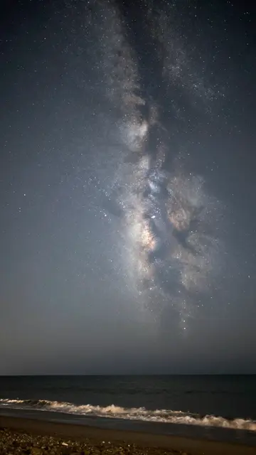 The glowing Milky Way galaxy stretches across a dark night sky above gentle surf on a quiet pebbled beach.