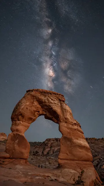 A natural sandstone arch stands beneath a vivid starry Milky Way night sky in a desert landscape