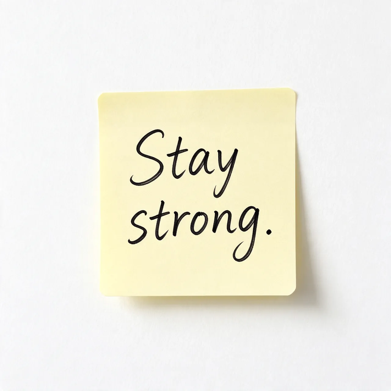 Handwritten "Stay strong." in black ink on a pale yellow sticky note against a plain white wall