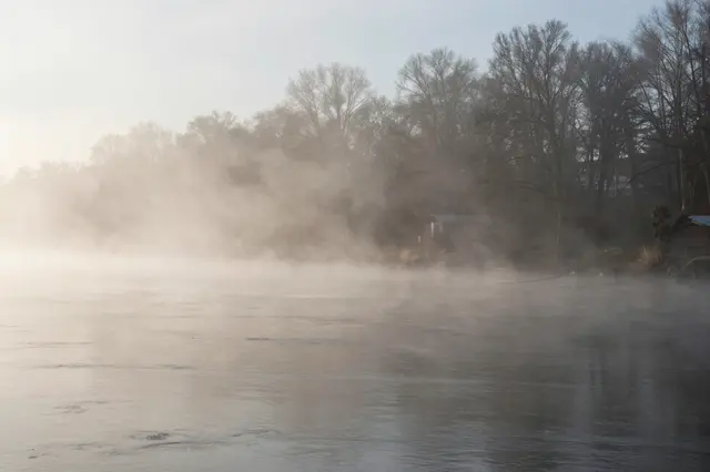 Thick steam fog rises from a river along a treed shore on a cold quiet morning