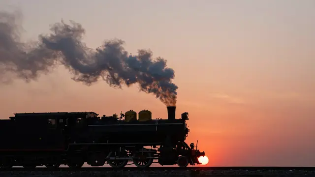 A steam train silhouette trailing wispy smoke passes in front of a glowing red sunset on the horizon
