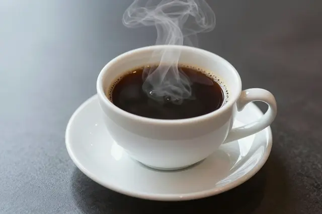 A white ceramic cup of hot black coffee with rising steam rests on a dark table surface.