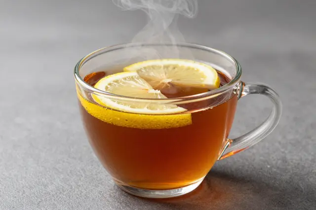 A clear glass cup holds hot black tea with lemon slices and wispy rising steam on a matte gray surface