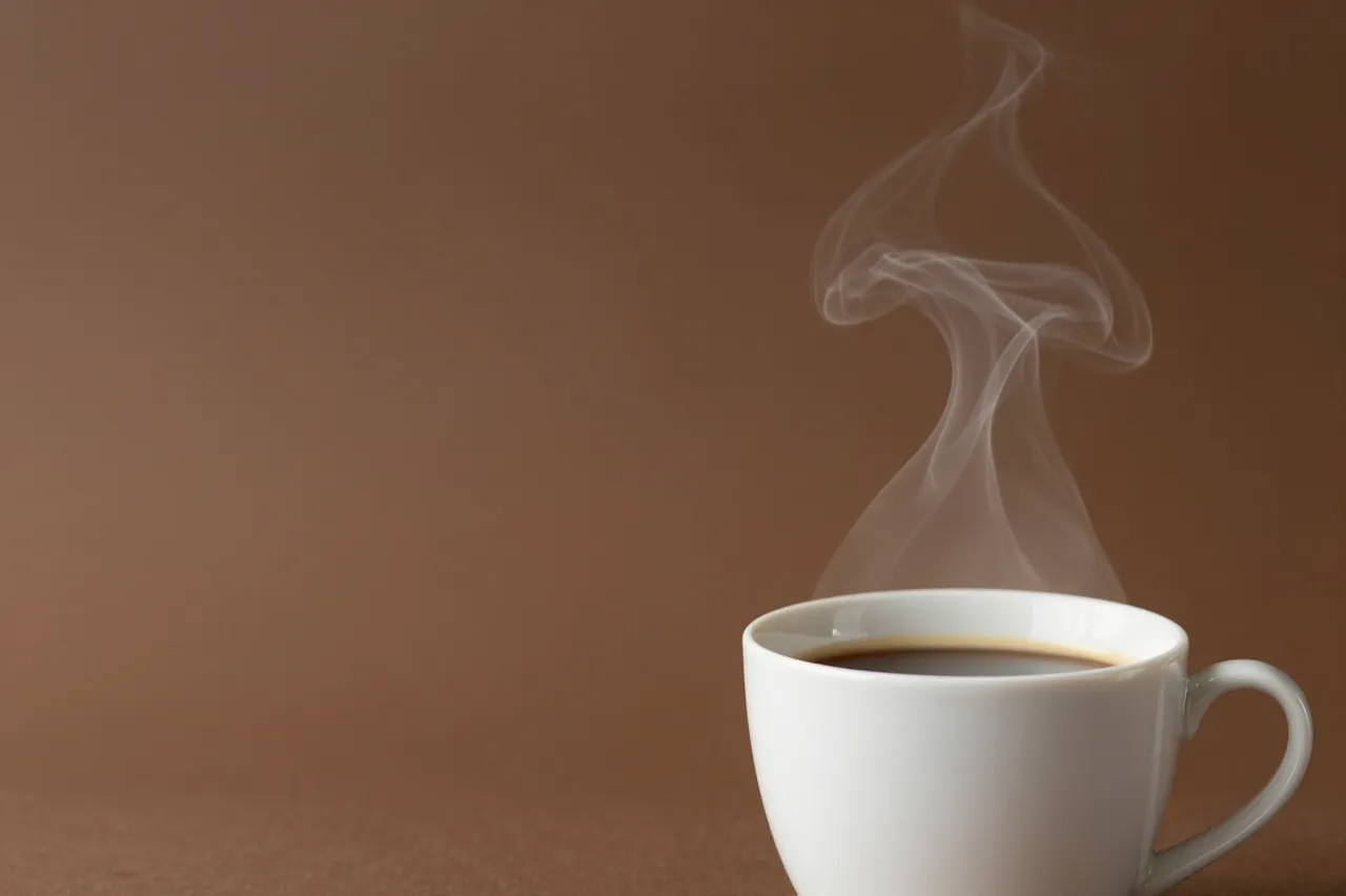 A white ceramic cup of hot coffee with rising steam sits at the bottom right of a warm solid brown backdrop with empty copy