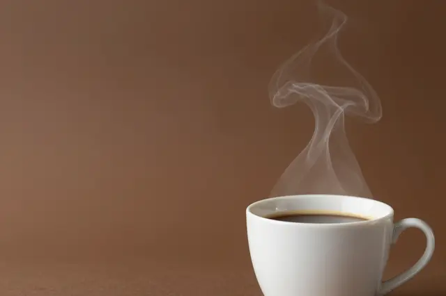 A white ceramic cup of hot coffee with rising steam sits at the bottom right of a warm solid brown backdrop with empty copy