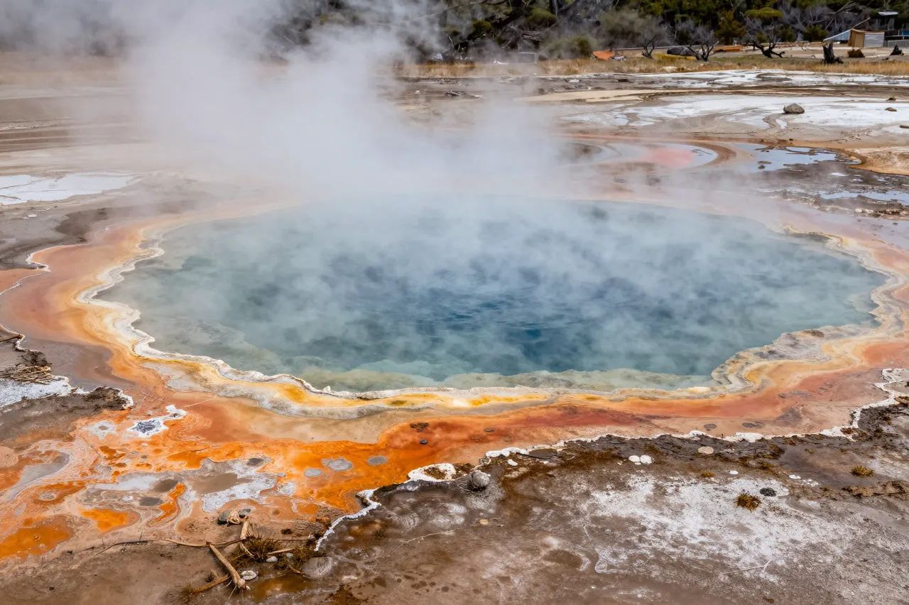 A colorful steaming hot spring with mineral rim bands sits in an open geyser basin landscape