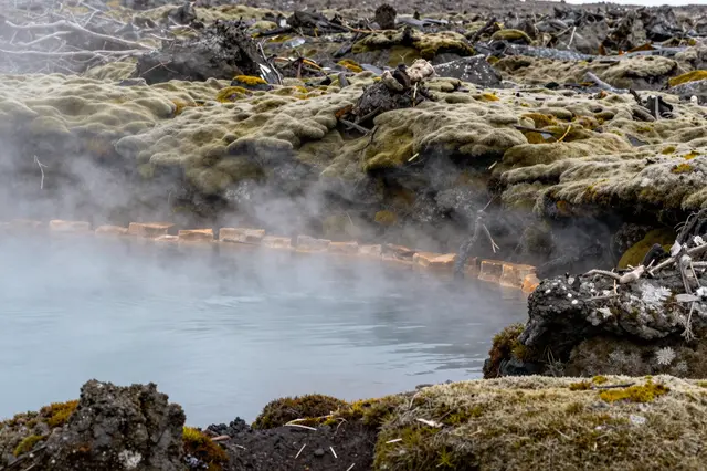 Wispy steam rises from a pale blue hot spring surrounded by thick moss-covered volcanic lava rock.