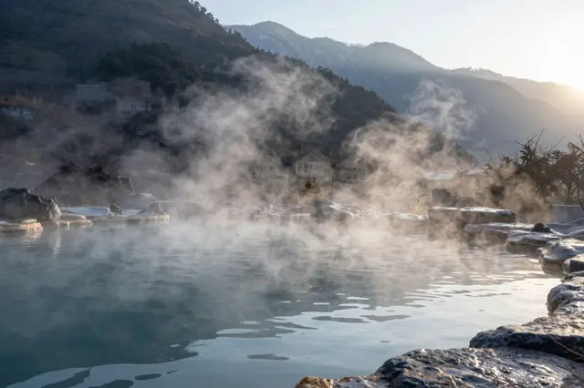 Steam rises from a mountain hot spring pool as golden sunrise light hits distant forested peaks
