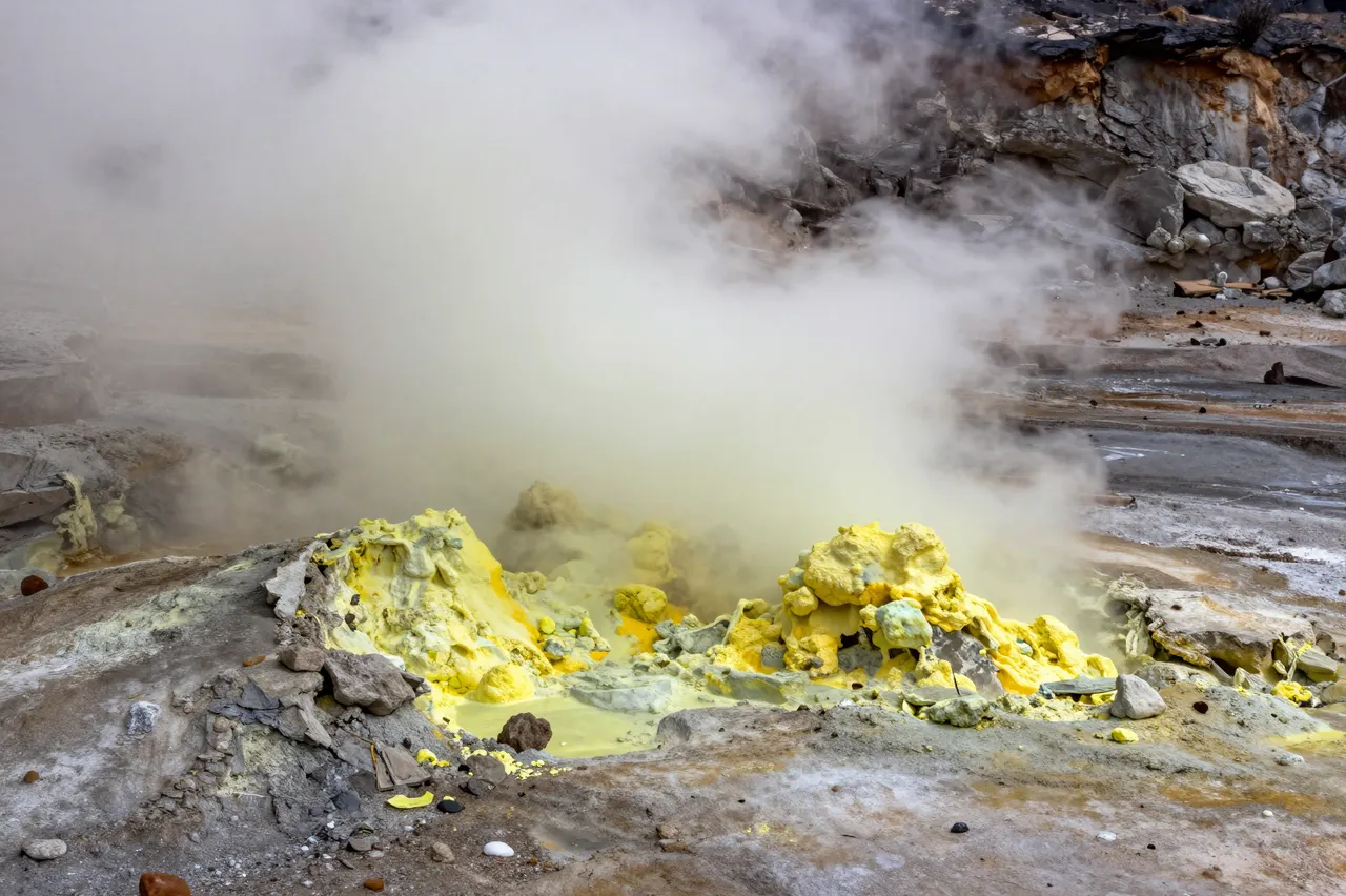 Thick steam rises from a volcanic crater lined with bright yellow sulfur deposits and rocky terrain