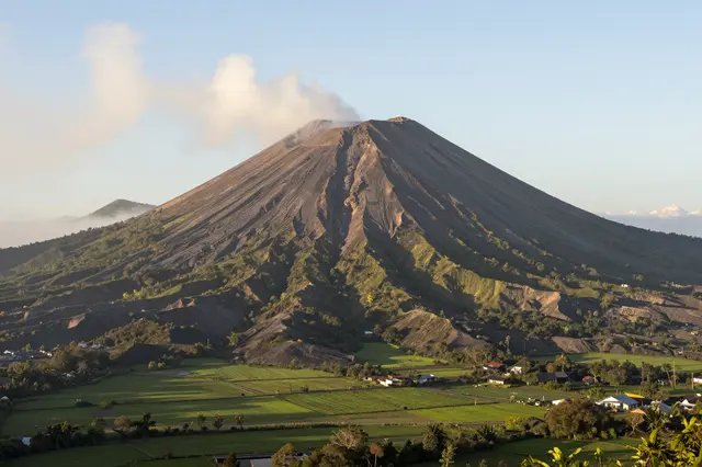 A gently steaming conical volcano rises above lush green farmland and small settlements at sunrise