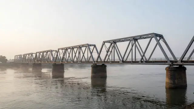 Empty steel truss bridge spans a misty wide river under soft hazy dawn light