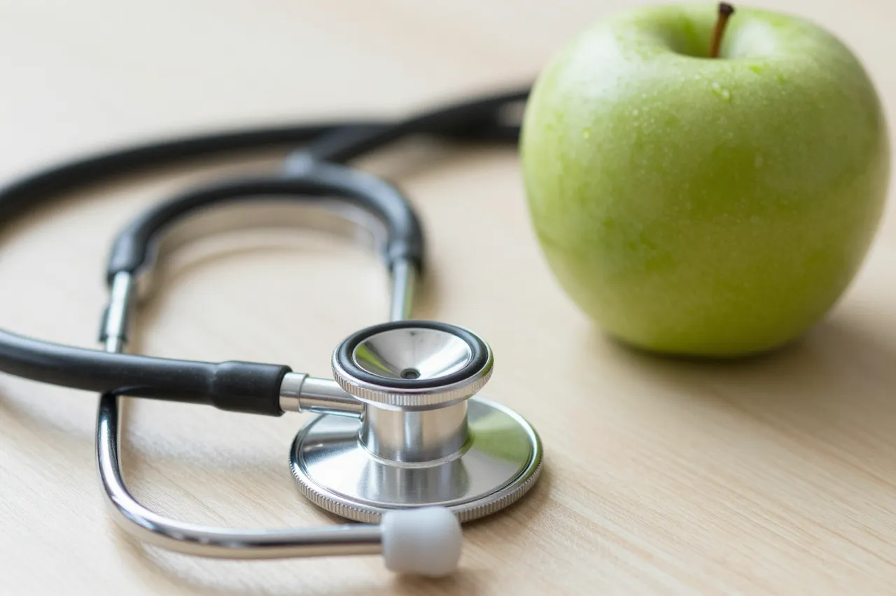 A stethoscope sits next to a fresh green apple on a light wooden desk surface