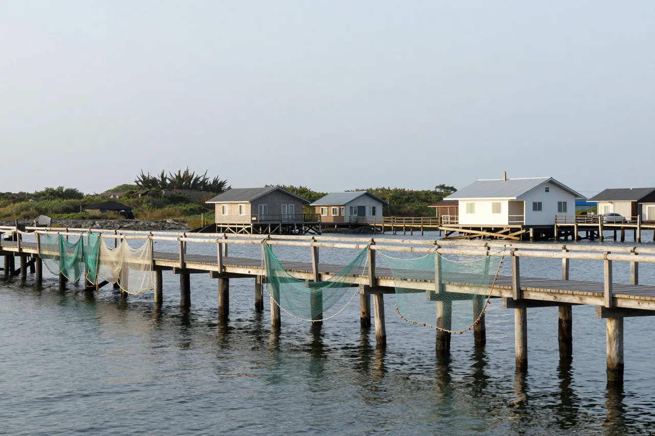 Fishing nets hang to dry on a wooden jetty beside stilt cottages under an overcast sky