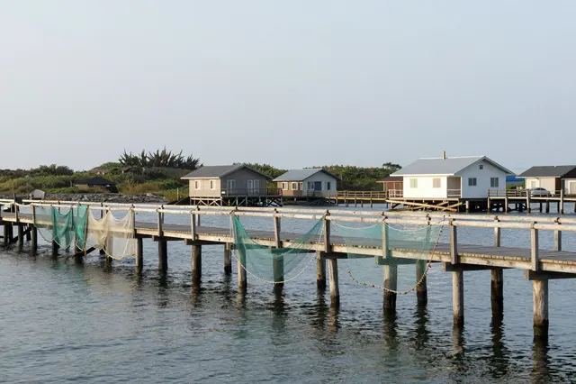 Fishing nets hang to dry on a wooden jetty beside stilt cottages under an overcast sky