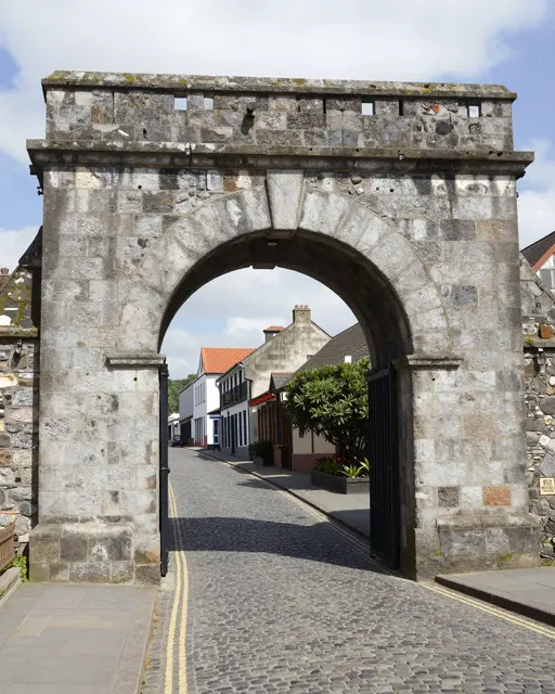Historic stone gate archway frames a sunny cobbled street lined with small buildings