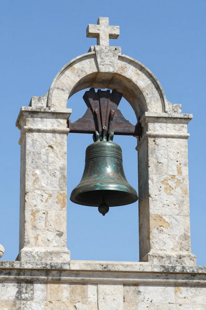 A patinated bronze church bell hangs in a weathered stone belfry against a clear bright blue sky.