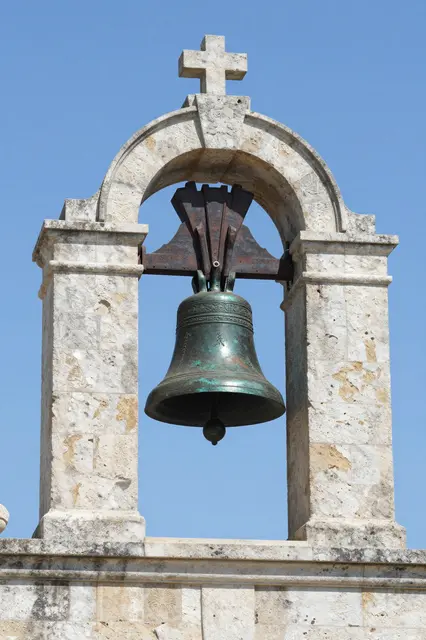A patinated bronze church bell hangs in a weathered stone belfry against a clear bright blue sky.