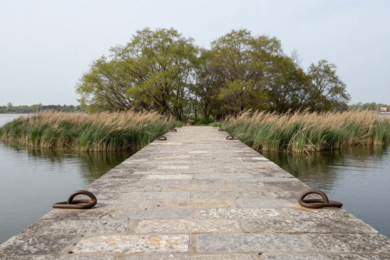Weathered stone jetty with iron mooring rings extends across calm water to a grassy tree-lined islet under a gray sky