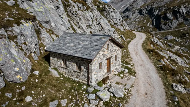 Aerial view of a rustic stone mountain hut beside a dirt trail on a high rocky alpine pass