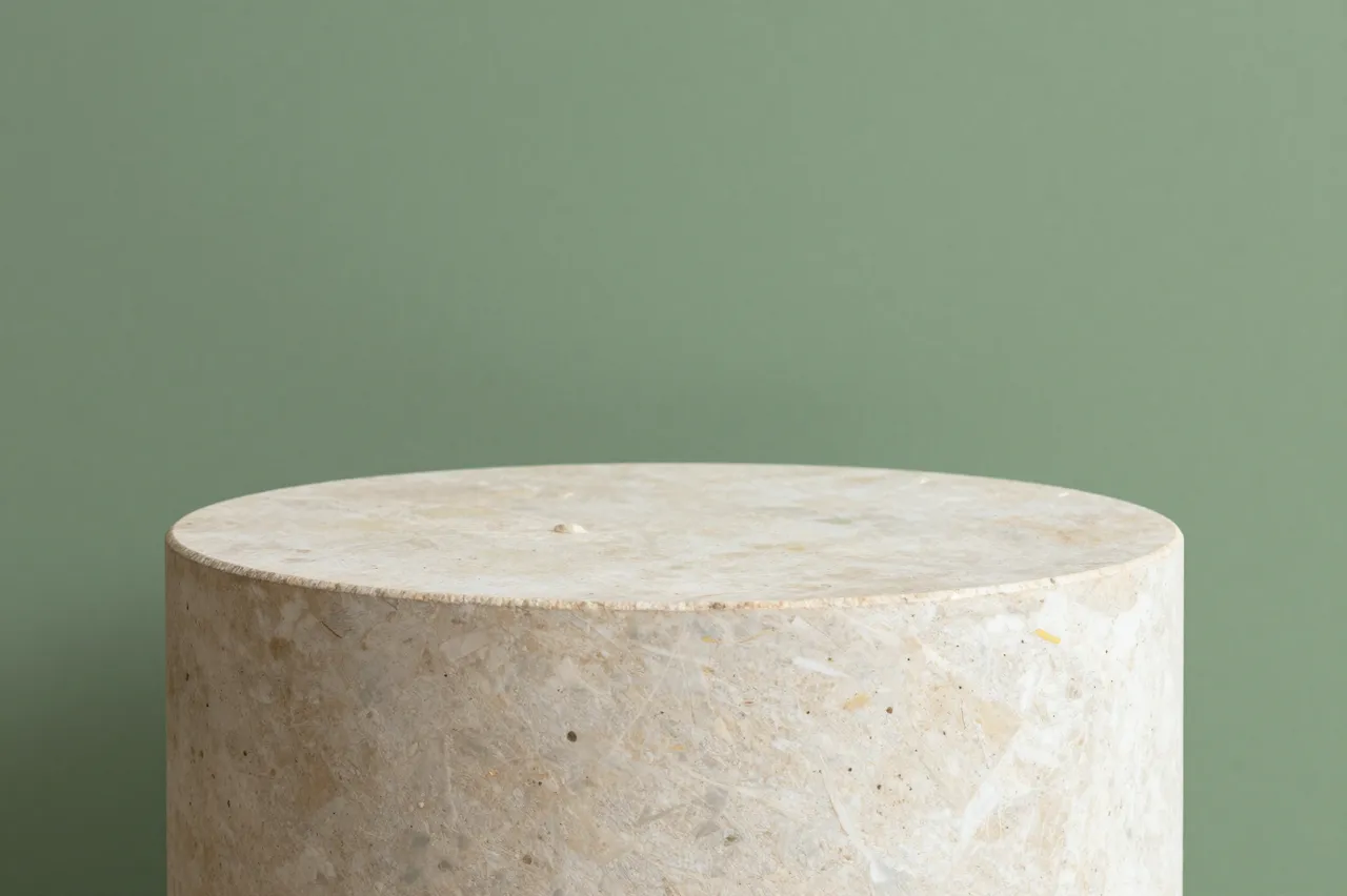 A cylindrical light stone product pedestal sits against a solid sage green wall