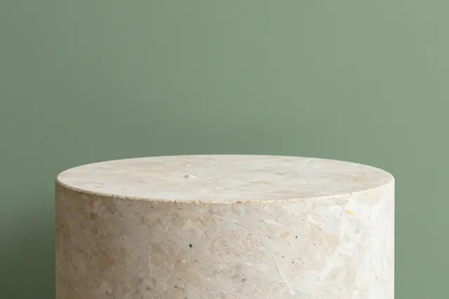A cylindrical light stone product pedestal sits against a solid sage green wall