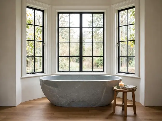 Freestanding gray stone bathtub sits beneath a black-framed bay window next to a wooden side table with a lit candle and