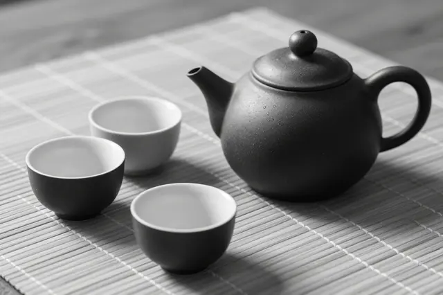 Black and white still life of a dark stoneware teapot with three small cups resting on a woven bamboo mat