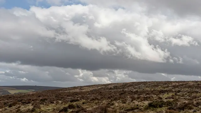 Turbulent wind-sculpted clouds drift across the sky above a wide open heather moorland ridge