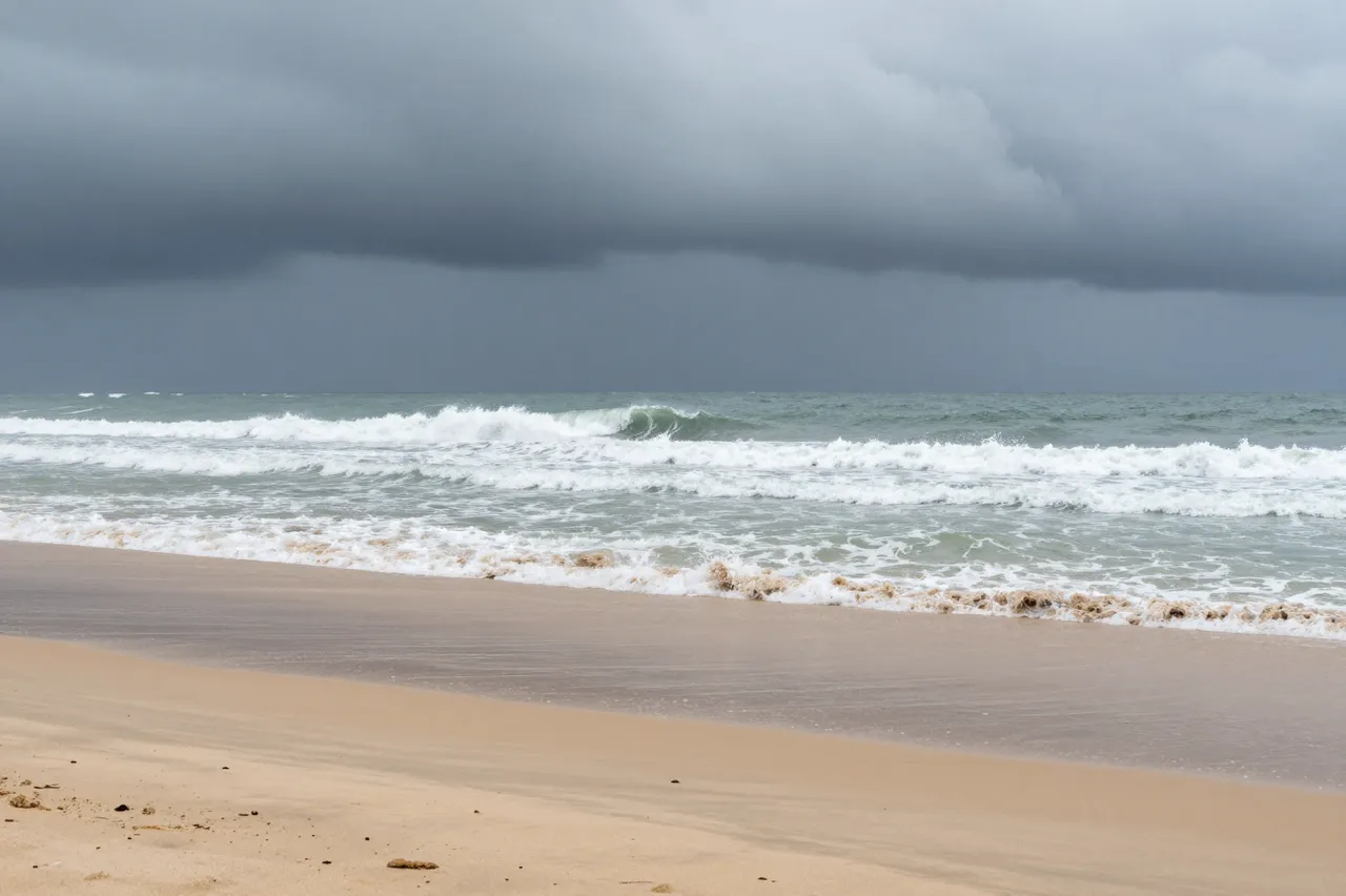 Dark heavy storm clouds loom above churning surf rolling onto a quiet empty sandy beach.