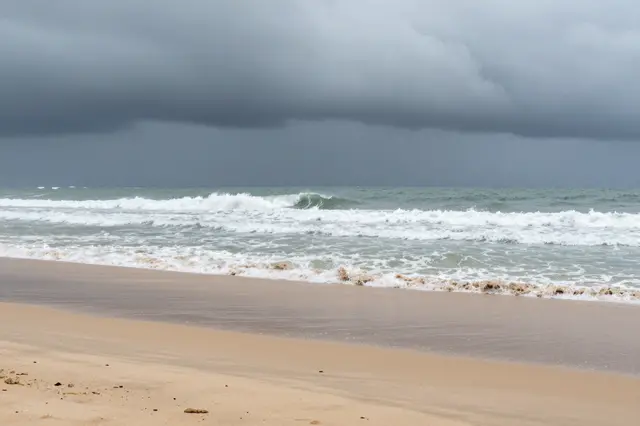 Dark heavy storm clouds loom above churning surf rolling onto a quiet empty sandy beach.