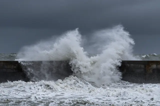 Massive foamy storm waves crash against a concrete harbor wall under a dark overcast sky