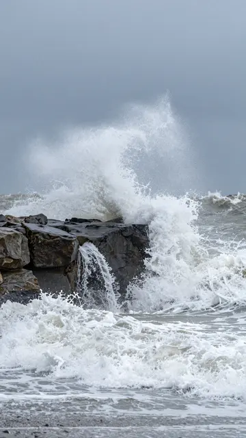 Turbulent storm waves crash and spray over dark coastal rock under an overcast slate sky