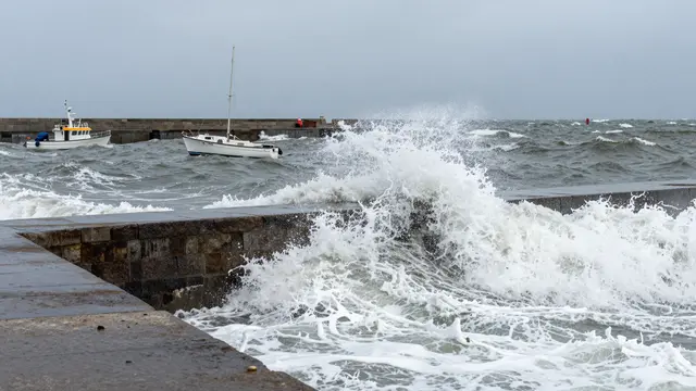 Waves crash over a stone harbor breakwater as small boats toss in choppy gray storm water