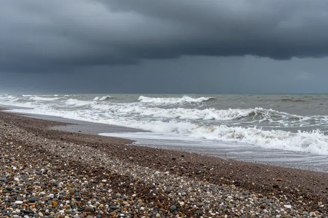 Turbulent foamy waves roll onto a multicolored pebble beach beneath heavy dark storm clouds.
