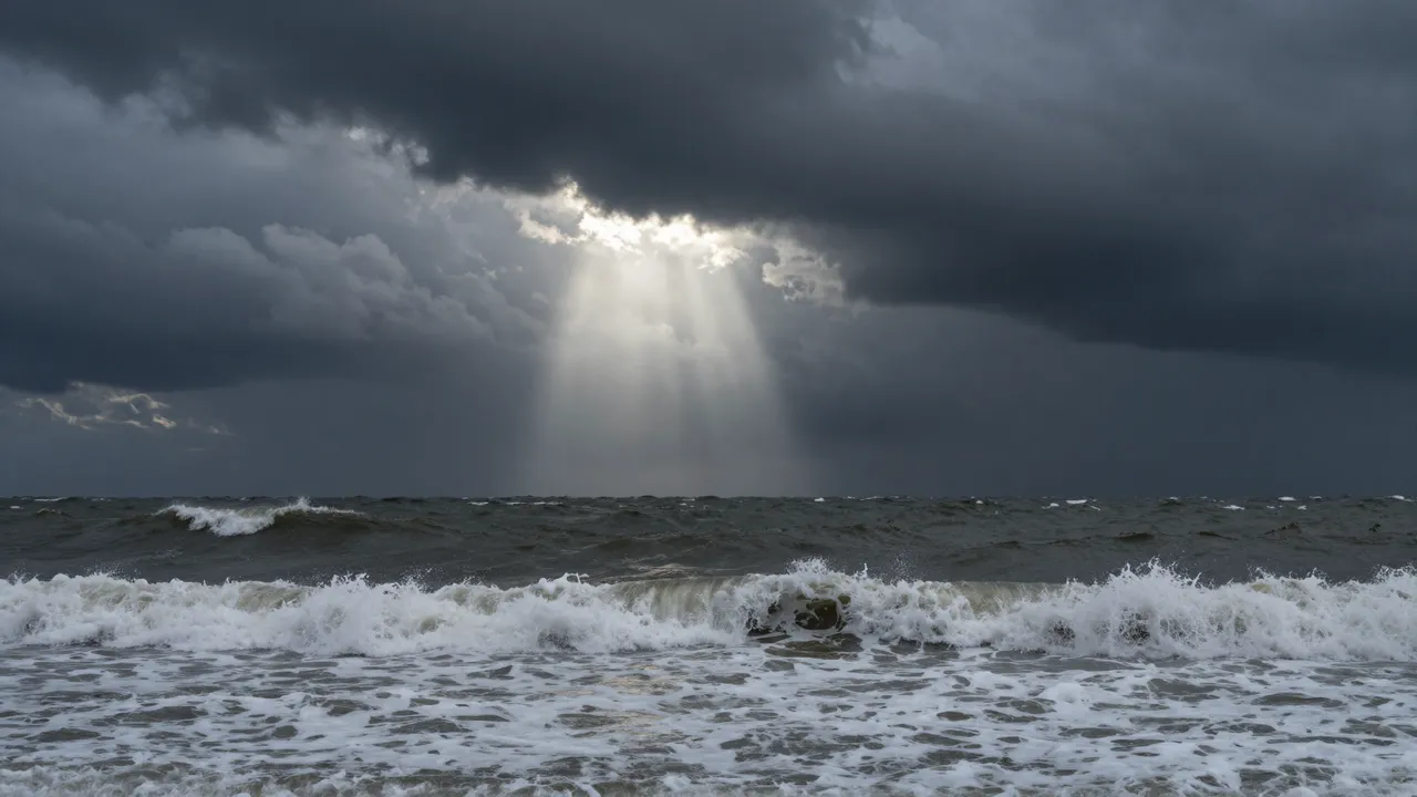 A single shaft of light cuts through dark storm clouds above churning, wave-tossed ocean swells.