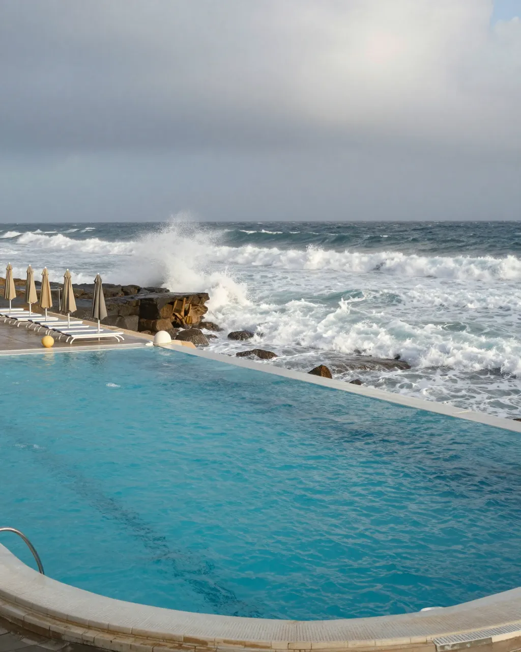 Infinity resort pool sits beside churning storm waves crashing on coastal rocks under overcast skies