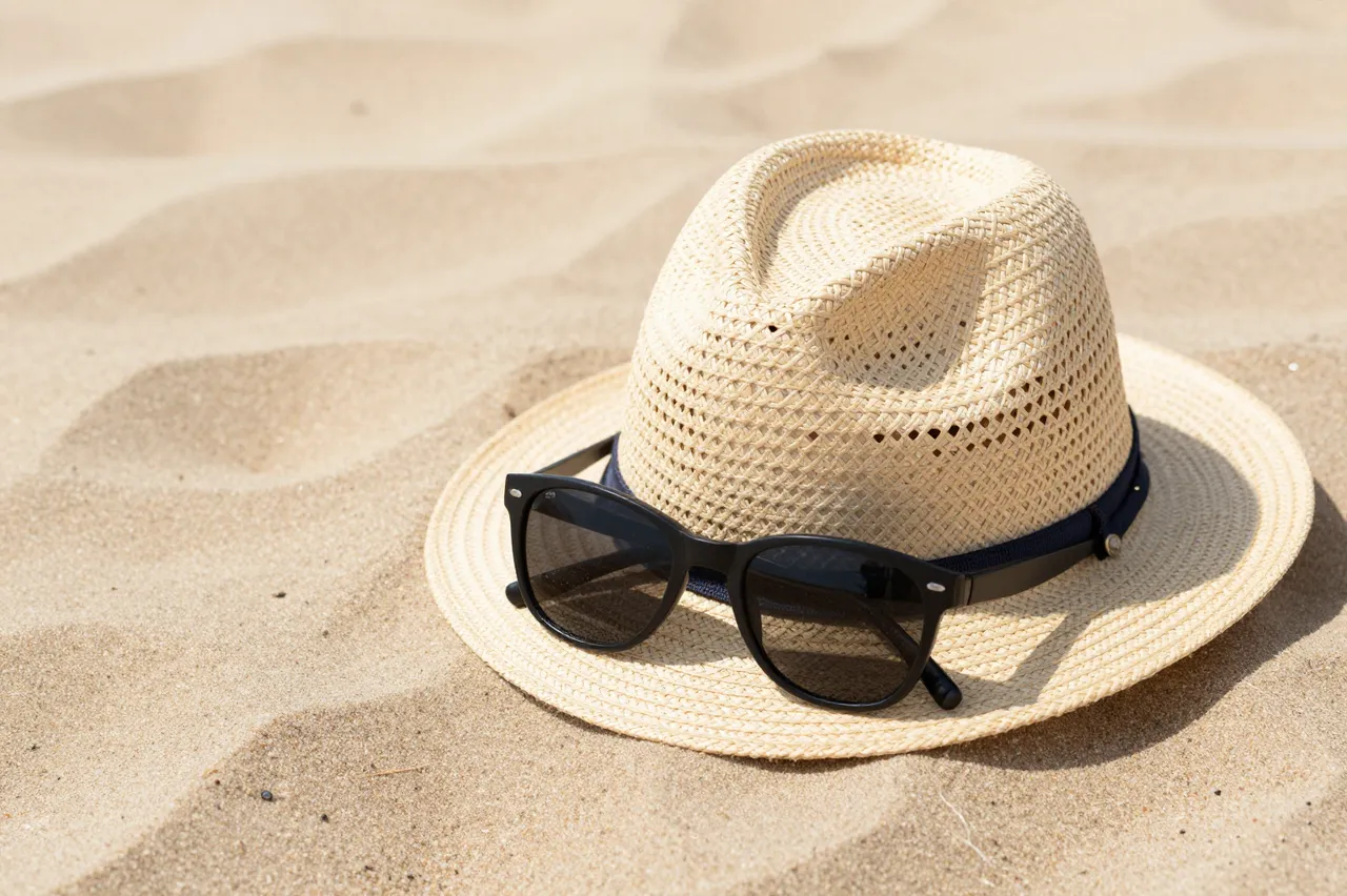 A woven straw hat with black sunglasses resting on its brim sits on rippled sunlit beach sand