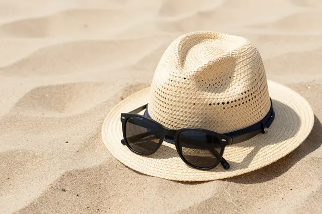 A woven straw hat with black sunglasses resting on its brim sits on rippled sunlit beach sand