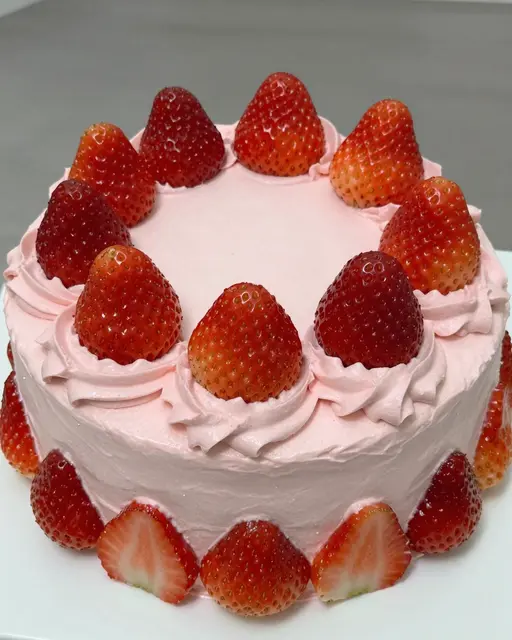 A round cake covered in light pink buttercream, decorated with whole and sliced fresh strawberries.
