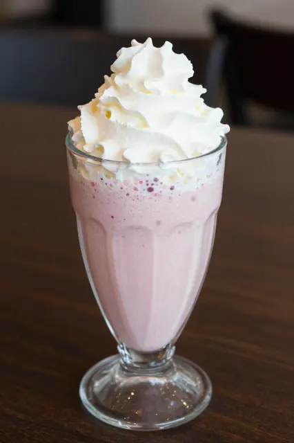 A tall glass of pink strawberry milkshake topped with piped whipped cream sits on a dark wood table.