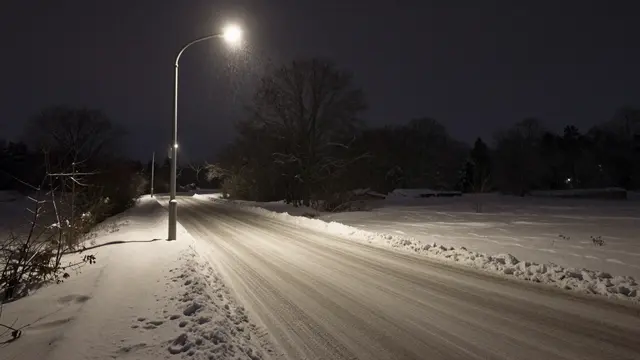 A lone glowing streetlamp illuminates an empty snow covered country road during a light snowfall at night
