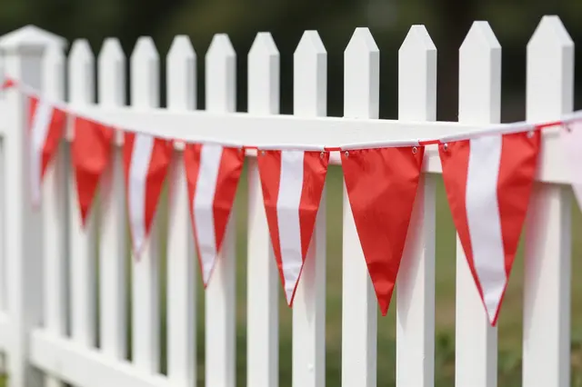 Red and white triangular striped bunting hangs draped along a white wooden picket fence in a sunny summer yard.