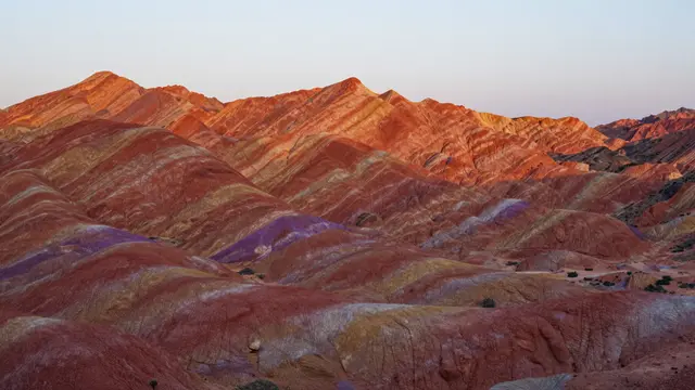 Layered desert mountains with red and purple geological bands glow under a soft pale sunset sky