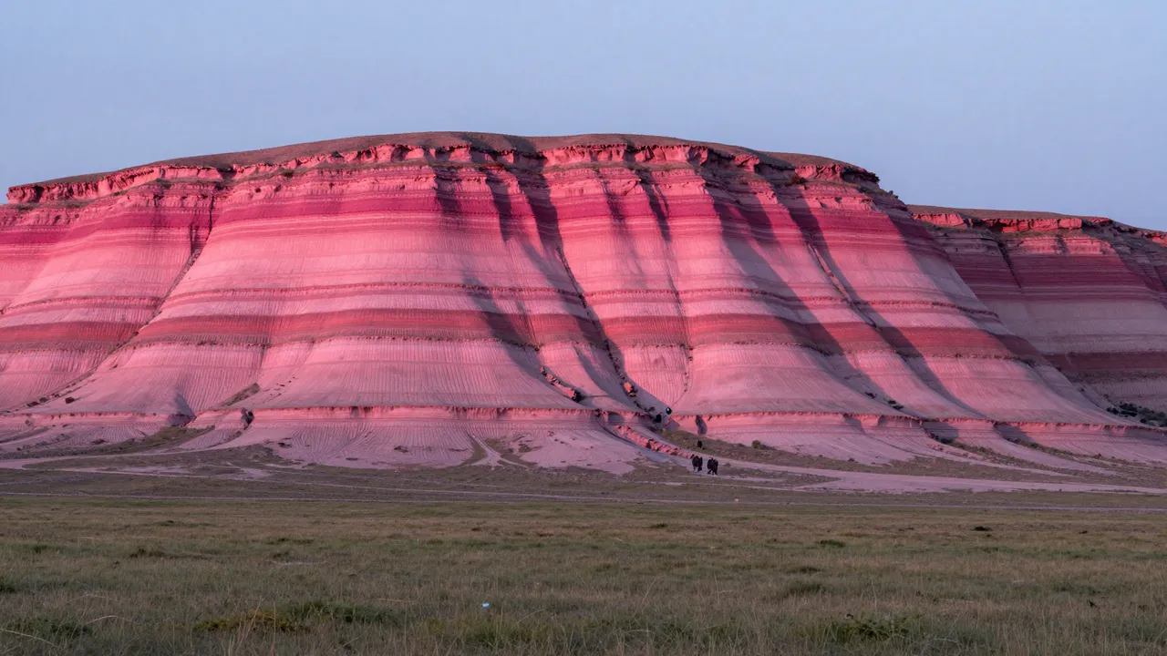 Layered striated pink mesa cliffs rise above flat grassy plain under a pale dusk sky