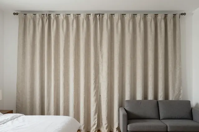 A linen ceiling track curtain separates a bed with white linens from a small gray sofa in a bright studio apartment.