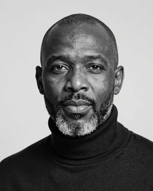 Black and white studio headshot of a middle aged Black man with a shaved head, salt-and-pepper beard, and dark turtleneck