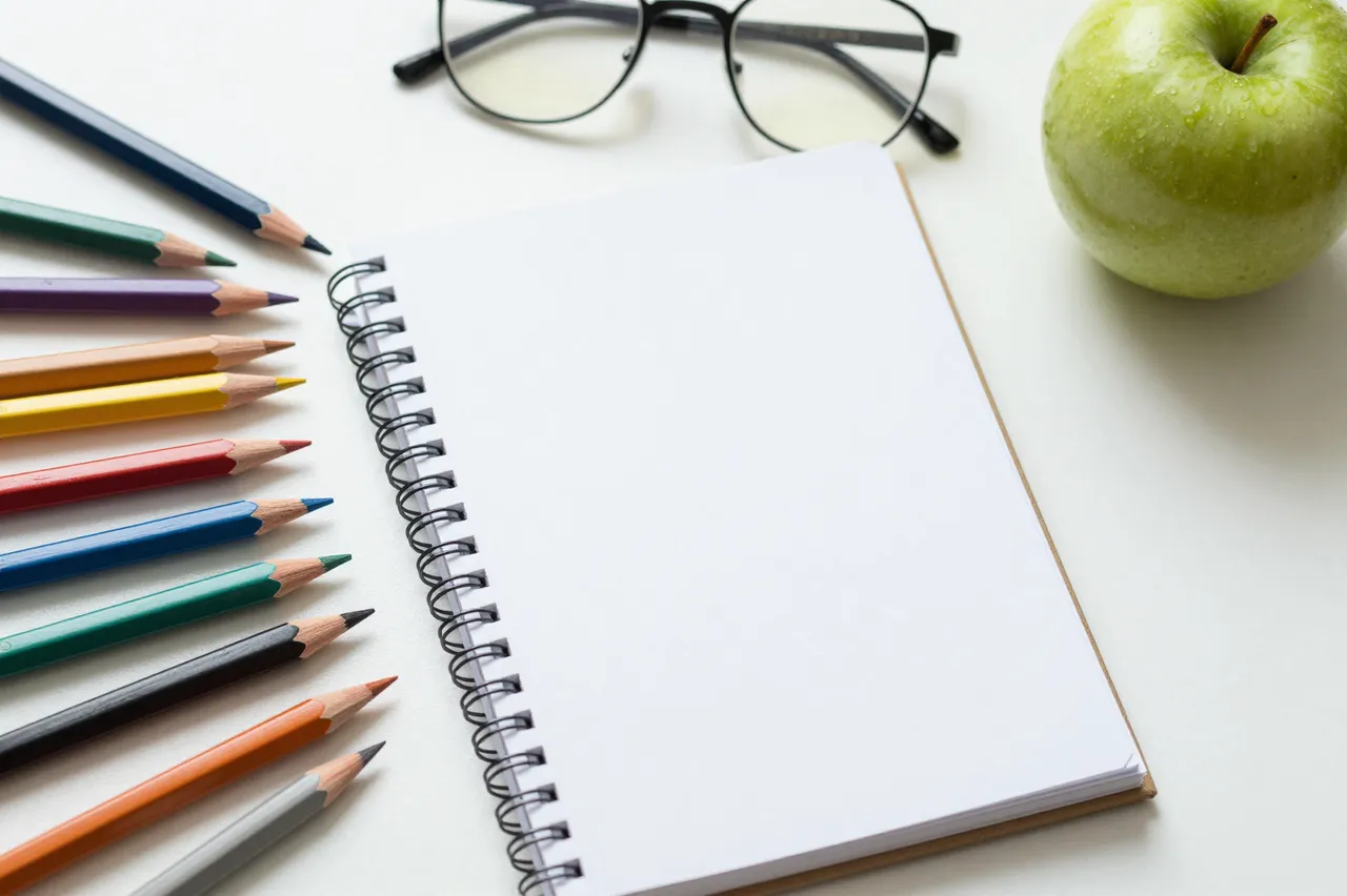 Colored pencils, blank spiral notebook, eyeglasses, and a green apple sit on a white desk surface