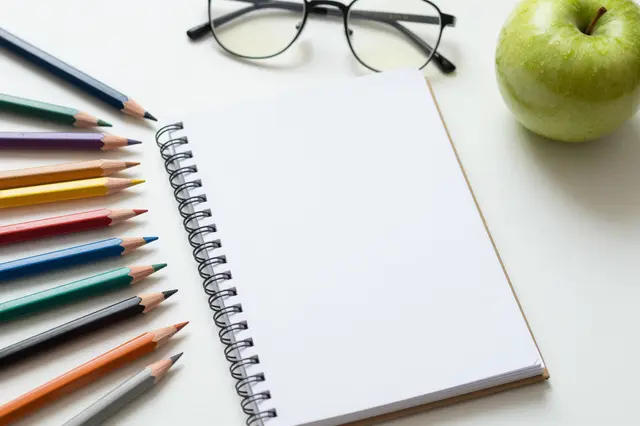 Colored pencils, blank spiral notebook, eyeglasses, and a green apple sit on a white desk surface