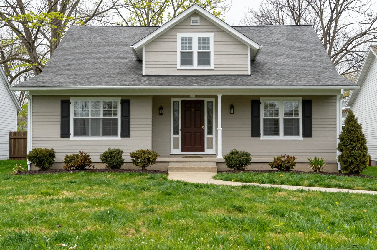 A light beige suburban house with a covered front porch sits above a green spring lawn between neighboring homes