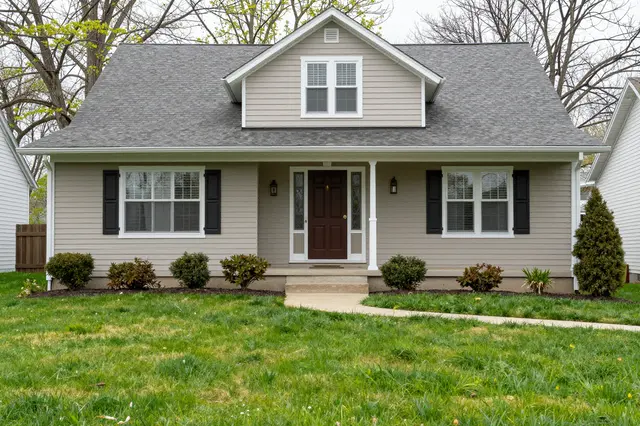 A light beige suburban house with a covered front porch sits above a green spring lawn between neighboring homes